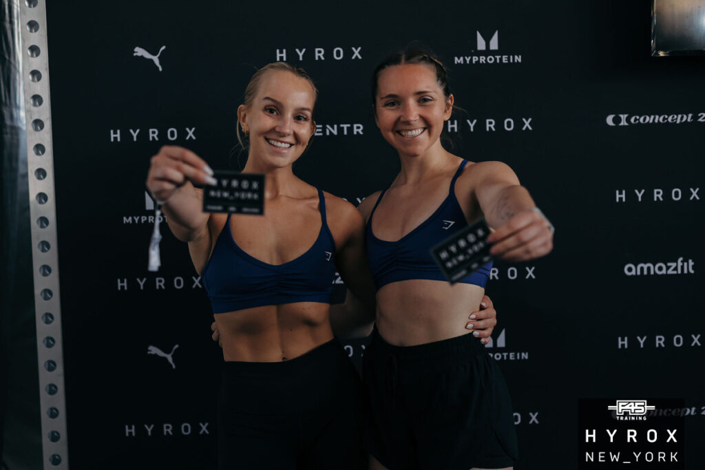 Two HYROX athletes smiling post-race while holding finish line tokens, standing in front of sponsor backdrop featuring HYROX, PUMA, and ERIMA logos