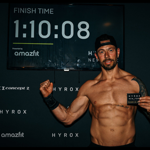 HYROX competitor flexes post-race in front of a results screen showing a finish time of 1:10:08, surrounded by fitness sponsor logos