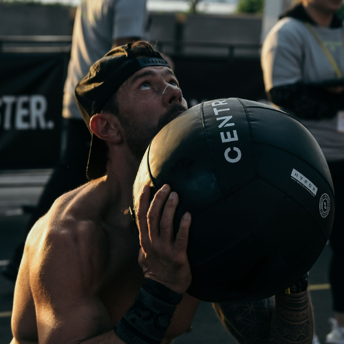 Athlete performing wall ball reps using a CENTR-branded medicine ball during HYROX training, with spectators and MONSTER banner in background.